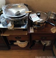 Cookware items including pans and wok stacked on wooden drawers, some wrapped in plastic or in box.