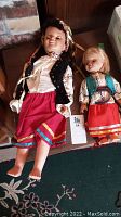 Two Ukrainian dolls, one taller with dark hair and ribbons, other shorter with blonde hair, both dressed in traditional clothing, shown lying down.