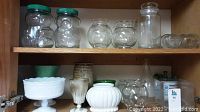 Shelf with two plastic peanut butter bear jars with green lids, small glass jars with scalloped edges, small round glass jars, tall cylindrical glass jar, white ceramic vases and others.