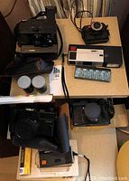 Overview of 6 vintage film cameras on wooden surface showing Polaroid OneStep, Akira 2000N, film canisters, flash unit, and other cameras