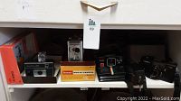 Front view of shelf with 5 vintage cameras including Kodak Brownie Movie Camera in box, Polaroid, Yashica, Halina, and Sanwa cameras.