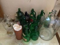 Wide view showing 13 large green wine bottles and 1 clear glass container on the floor near a wall.