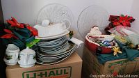 Wide view of stacked Christmas dinnerware including Grindley set, glass serving plates, cloth napkins with poinsettia rings, and ceramic bowls placed on boxes