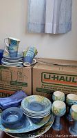 Assorted hand painted plates, bowls, and mugs stacked on moving boxes.