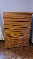 Tall boy dresser showing five drawers with large wood handle pulls, wood finish, standing on wood flooring.