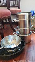 Stack of four stainless steel pots including a double boiler, with a metal strainer and black enamel roaster beneath.