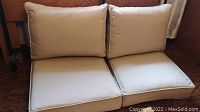 Two beige seat and back cushions stacked and set on wooden furniture showing upholstery and dimensions.