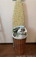 Photo of an ironing board with metal frame and patterned cover standing upright behind a lined wicker hamper topped with Black & Decker iron, fabric steamer, and instruction manual.