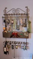Metal wall-mounted jewelry organizer with hanging costume necklaces, bracelets, earrings, and a small shelf box area.