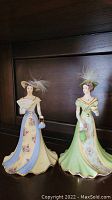 Two Lena Liu porcelain figurines placed side by side on a wooden shelf, showing full frontal and side angles of dresses, hats, and accessories.