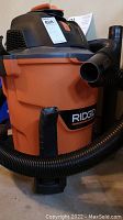 Front view of the orange and black Ridgid Wet Dry Vac HD12000, showing hose attached and the body with Ridgid branding.
