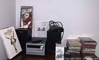 Panasonic stereo, Hyundai mini speakers, DVD sets of Louis Armstrong and Michael Jackson, collection of CDs, and a Craig alarm clock displayed on a table.