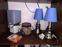Three mini lamps on shelf: two matching silver metallic base lamps with blue conical lampshades; one ceramic base lamp with square gray lampshade; and a Himalayan salt lamp with wax melting tray and scented wax supply.