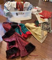 Photo showing clear plastic tote with various linens including holiday-themed tea towels, table runners, and garment-like pieces laid around it on wood floor.