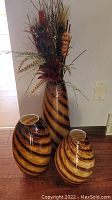 Full view of three painted glass vases on floor, tallest vase contains artificial foliage including cattails and peacock feather