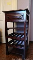 Front and side view of a dark wooden wine cabinet showing the top drawer with metal ring pulls, a middle shelf, and two slatted bottle racks below.