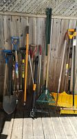 Group photo of around 17 lawn tools including spades, shovels, rakes, and brooms lined up against a wooden fence.
