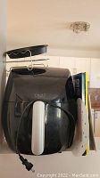 Front and side view of black Yedi Houseware air fryer showing white handle, power cord, metal rack on top, and open cookbook behind.