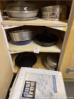 Shelving unit with multiple cookware items including baking pans, iron skillets, a casserole dish, and the dust-free cup and glass saver set in packaging in front