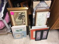 Photo showing multiple framed and unframed wall art pieces stacked and leaning against a wall, including tropical palm trees and botanical art with various frame styles and sizes