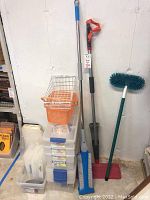 Photo of storage bins and cleaning tools arranged near a wall including mops, brushes, and plastic bins.