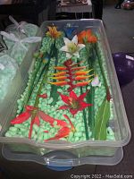 Plastic container filled with green packing peanuts holding a set of wood painted decorative flowers including various colors and types.