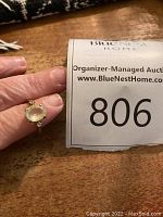 Photo showing the 10K gold ring with an oval yellow-orange center stone and small clear accent stones, held in hand next to auction number.