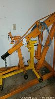 Side view of orange steel hydraulic shop crane with coiled metal strap on boom and yellow engine stand visible