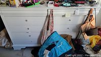 Photo of a white wooden dresser with six drawers and round knobs, showing scratches. Clothing and bags are placed around and hanging from the dresser. Contents on dresser not included.