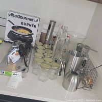 Wide shot showing Elite Gourmet electric burner box, glass jars with lids, bottles, barware including shaker, jigger, pepper grinder, tumblers, and flatware on countertop