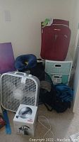 Photo showing white box fan, red suitcases stacked on teal drawer organizer, blue neck pillow, and a blue backpack on floor.
