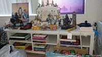 Altar setup with various Hindu and Buddha statues, framed picture, books below altar in cubby spaces, prayer beads and incense holders visible