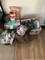 Wide view showing Aero Garden system box, multiple colorful plant pots stacked, woven picnic basket with handles, aprons with cherry pattern, plastic cutlery and napkins in plastic packaging, and other gardening supplies.