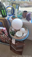 Full view of side table topped with a cream tray holding decorative balls, green perfume bottle, and small bowls. Surrounding items include framed artwork, glass objects, and crystals captured in natural light.