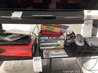 Sony DVD player DVP SRS10H on original box with remote on top, and nearby Super Nintendo style TV game console with wired controller, along with a stack of DVDs and VHS tapes.