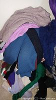 Pile of assorted women's sweaters including beige cable knit, purple, blue ribbed, black ribbed, navy plush sweaters.