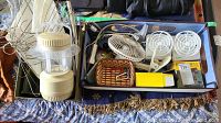 Overview of lot with beige battery-operated Dorcel lamp, blue storage container filled with various items including two small white desk fans, a woven basket with keys and accessories, and other bags and boxes visible around them.