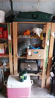 Camping items on wooden shelving including Martin propane BBQ, propane bottles, cooler, tent in bag, rope and other camping gear.