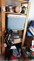 Wooden shelving unit holding blue Coleman cooler, stainless steel pots, insulated beverage container, propane bottles, red and black folding camping chair, other camping items piled underneath