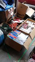 Boxes containing assorted Christmas decorations such as ribbons, garlands, extension cords, and boxed Christmas village houses.
