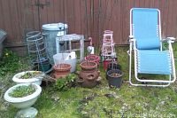 Wide view of two matching clay pots, white plastic plant pots, smaller black plant pots, several tomato cages, a solar light, a galvanized metal garbage can, and a blue folding lounger on grass near a wooden fence.