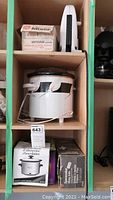 GE mixer, Rival Crock-Pot slow cooker, Better Crocker mini warmer, Snack Time sandwich maker on cabinet shelf with boxed items.