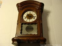 Full view of Franz Hermle wall clock with carved wooden case and glass front showing clock face and pendulum window.