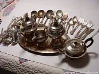 Photo of the full tea set and assorted silver plated cutlery laid out on a white surface showing teapot, sugar bowl, creamer, tray and various spoons and forks.