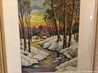 Full view of framed needlework showing winter landscape with snow, trees, houses, and sunset sky.
