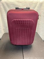 Front view of maroon VB Design carry on bag showing hard molded front panel with logo embossing and handle on top.