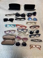 Full view of 17 pairs of eyeglasses and sunglasses arranged in rows on a white textured surface, including 2 open glasses cases.