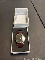 Larsson & Jennings wristwatch displayed in open white box with foam lining, showing the gold-tone case and burgundy leather strap.