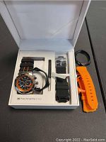 Open box showing TechnoMarine chronograph wristwatch with black and orange dial, orange and black silicone strap, two additional replacement straps (black and orange), black rubber strap holder, and tool for strap adjustment.