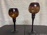Two decorative glass cups shown standing on a black surface with a textured white background, highlighting their tall stems and amber bubble-patterned bowls.